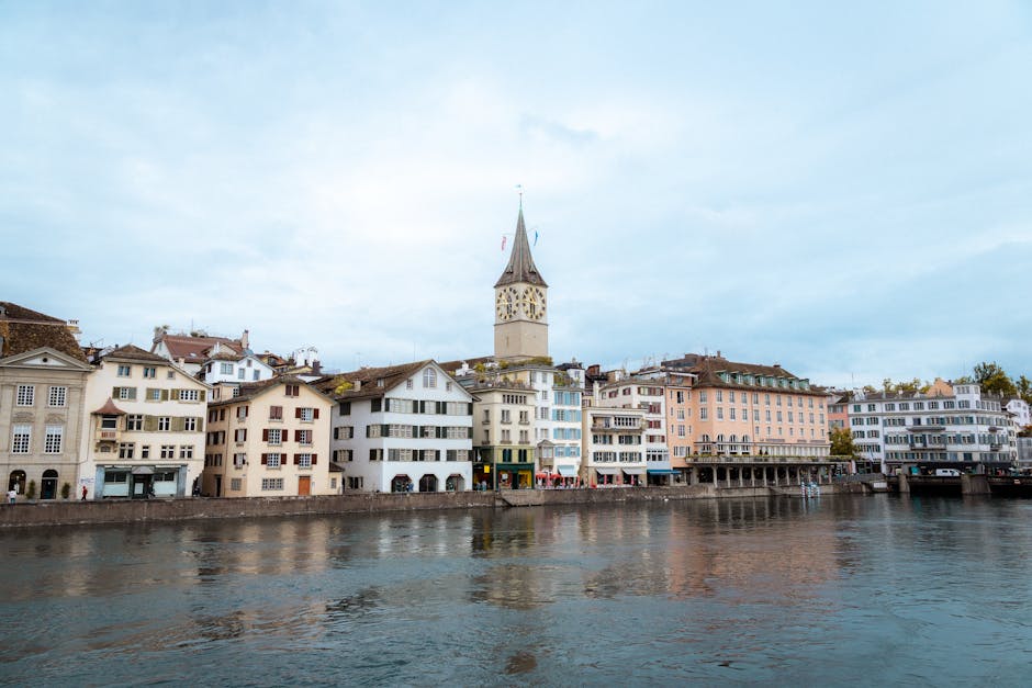 Scenic view of Zürich's historic buildings and clock tower by Lake Zürich under a cloudy sky.